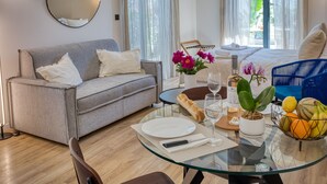 Habitación de hotel moderna con un sofá gris, una mesa de comedor de vidrio con platos, una botella de vino y una cesta de frutas.