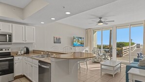 A modern kitchen with white cabinets, a built-in microwave, and a dishwasher. There is a dining area with a white table and chairs, and a large window offering a view of the outdoors. - Waters Edge 115 ~ Ground Floor ~ Beach Boardwalk Right Outside Your Door! (Fort Walton Beach)