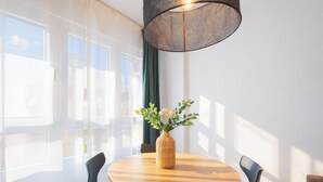 Ett rum med ett runt bord, stolar, en vas med blommor, tunna gardiner och en hängande lampa.