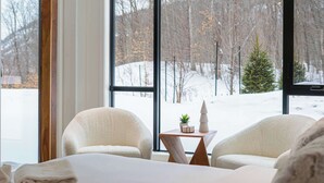 Ein gemütliches Zimmer mit großem Fenster, zwei Sesseln, einem kleinen Tisch mit Pflanze und verschneiter Aussicht.