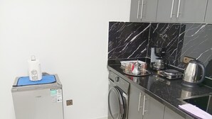 Cocina compacta con lavadora, cafetera y hervidor sobre la encimera.