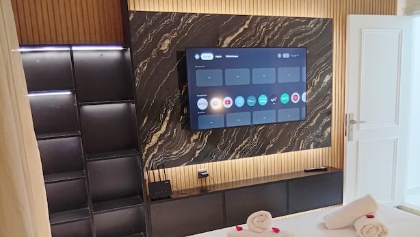 Una habitación de hotel moderna con un televisor de pantalla plana grande montado en una pared de mármol, una cama con sábanas blancas y pétalos de rosa, y un diseño de paneles de madera en la pared.
