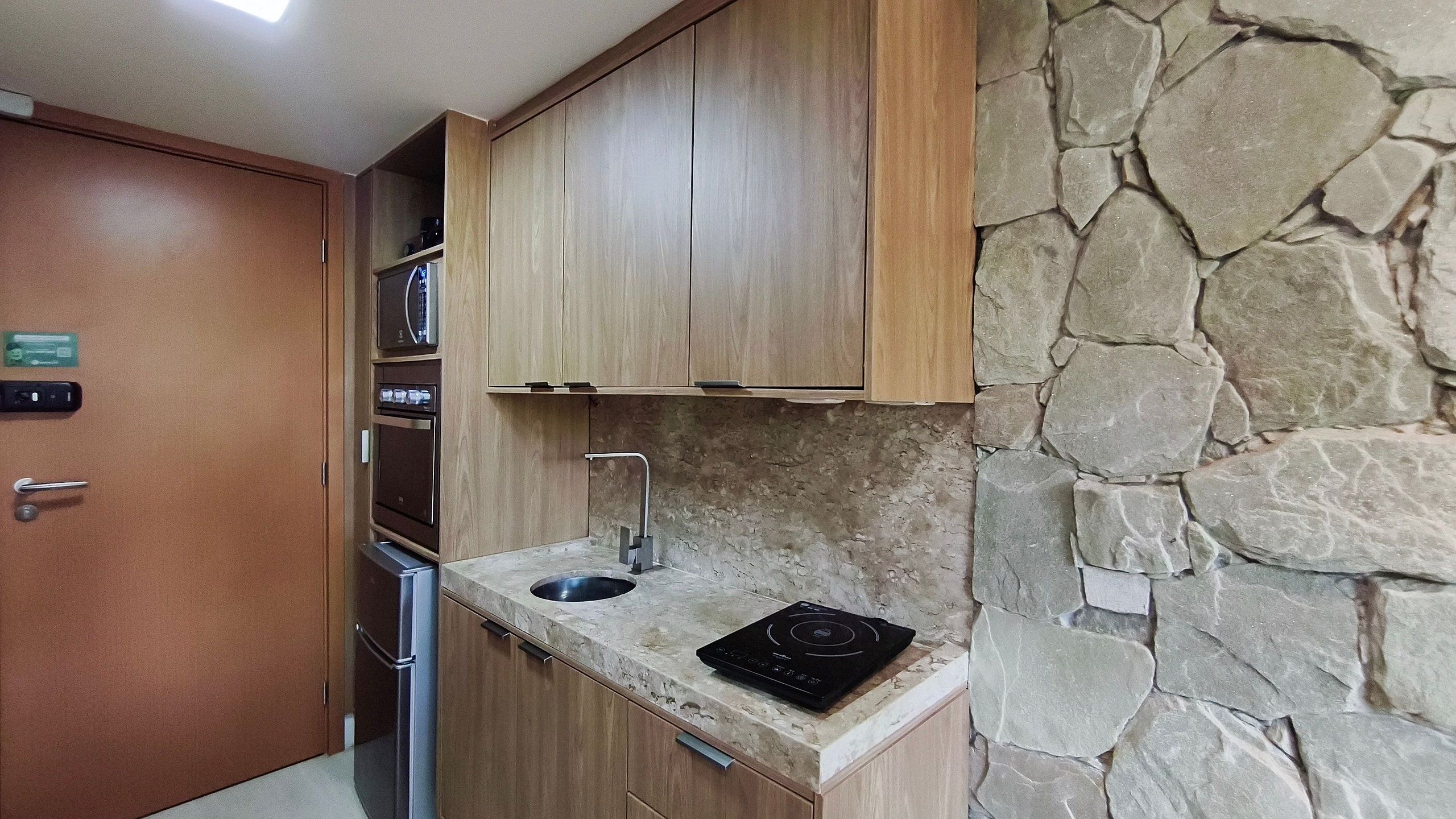 Une cuisine compacte avec des meubles en bois, un four encastré, un four à micro-ondes, un évier et un mur en pierre.