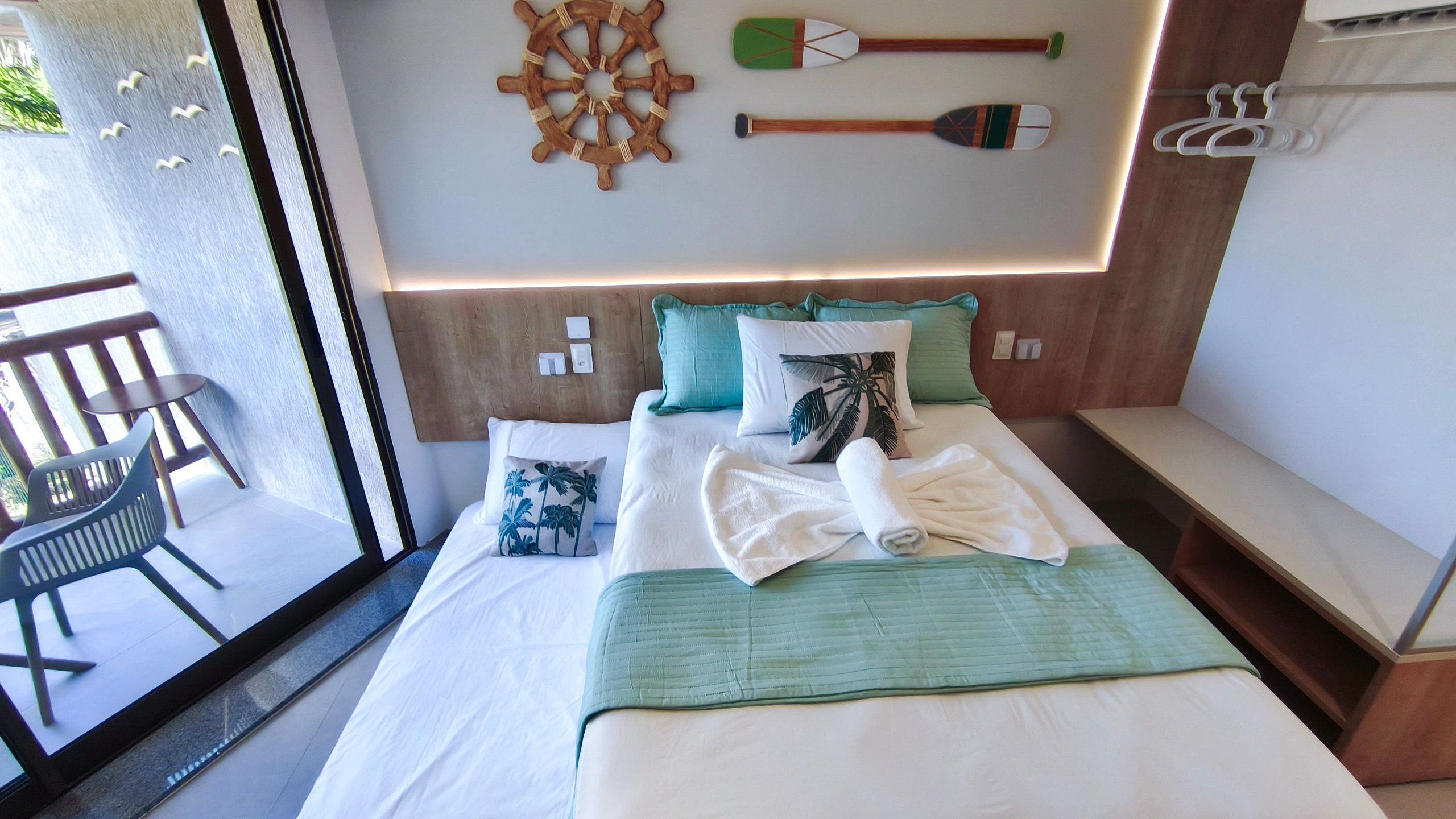 Quarto de hotel com cama, almofadas, toalha, cabeceira, remo fixado na parede e varanda com uma cadeira.