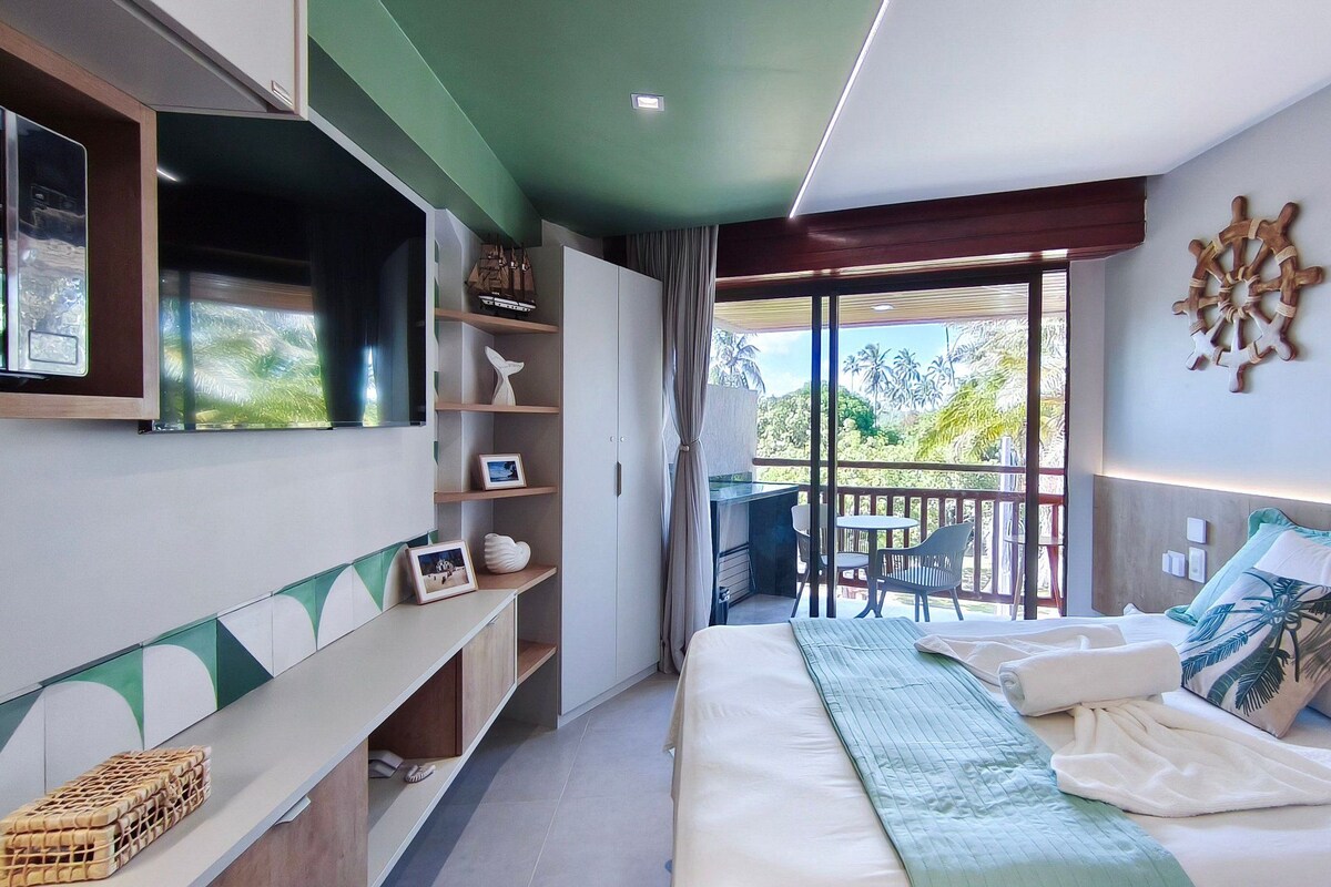 A modern hotel room with a balcony, a bed with a green quilt, a TV mounted on the wall, and a shelf with decorative items.