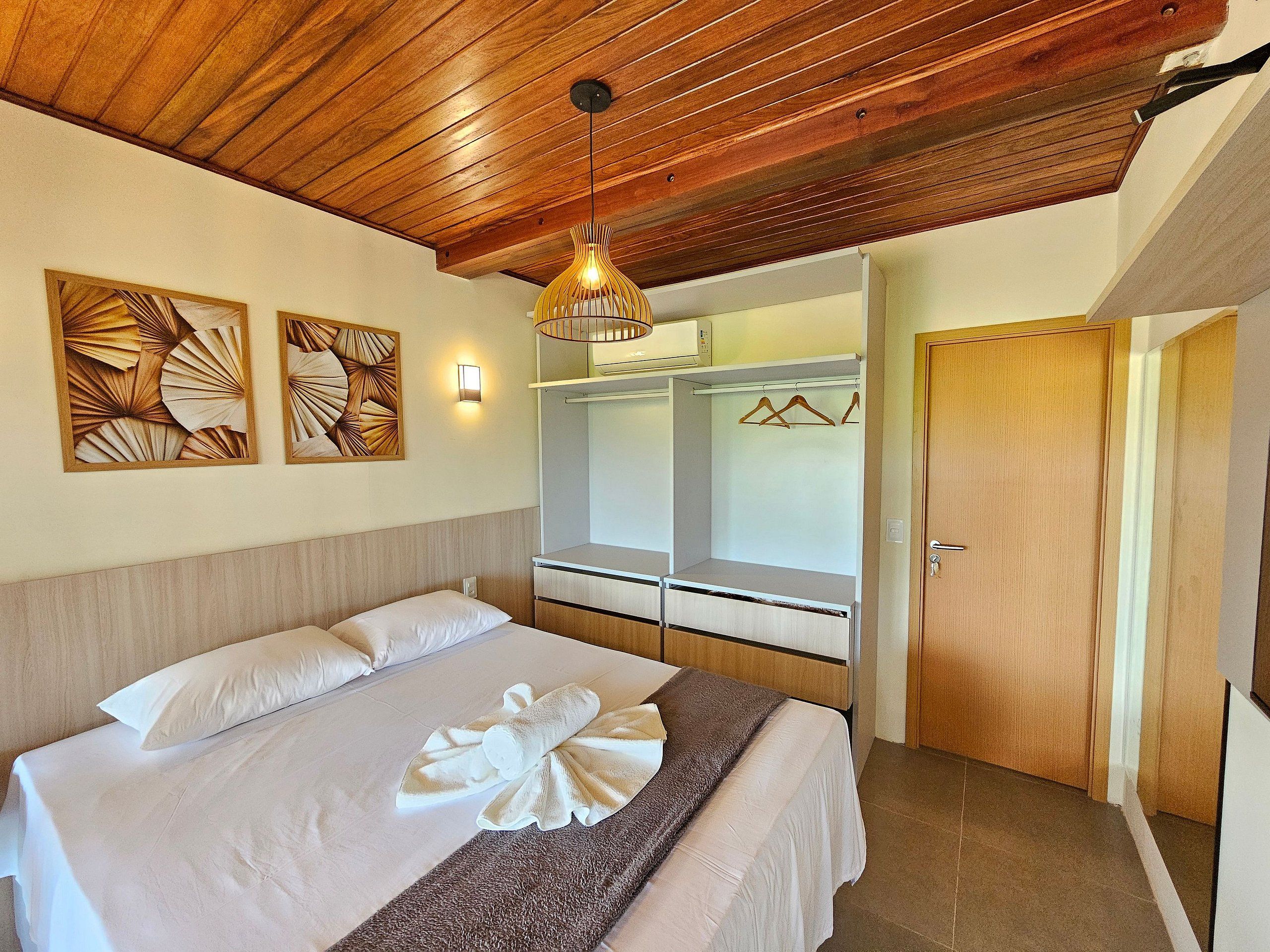 A bedroom with a wooden ceiling, a bed with white linens, a wall-mounted light, and a wardrobe with a mirror.