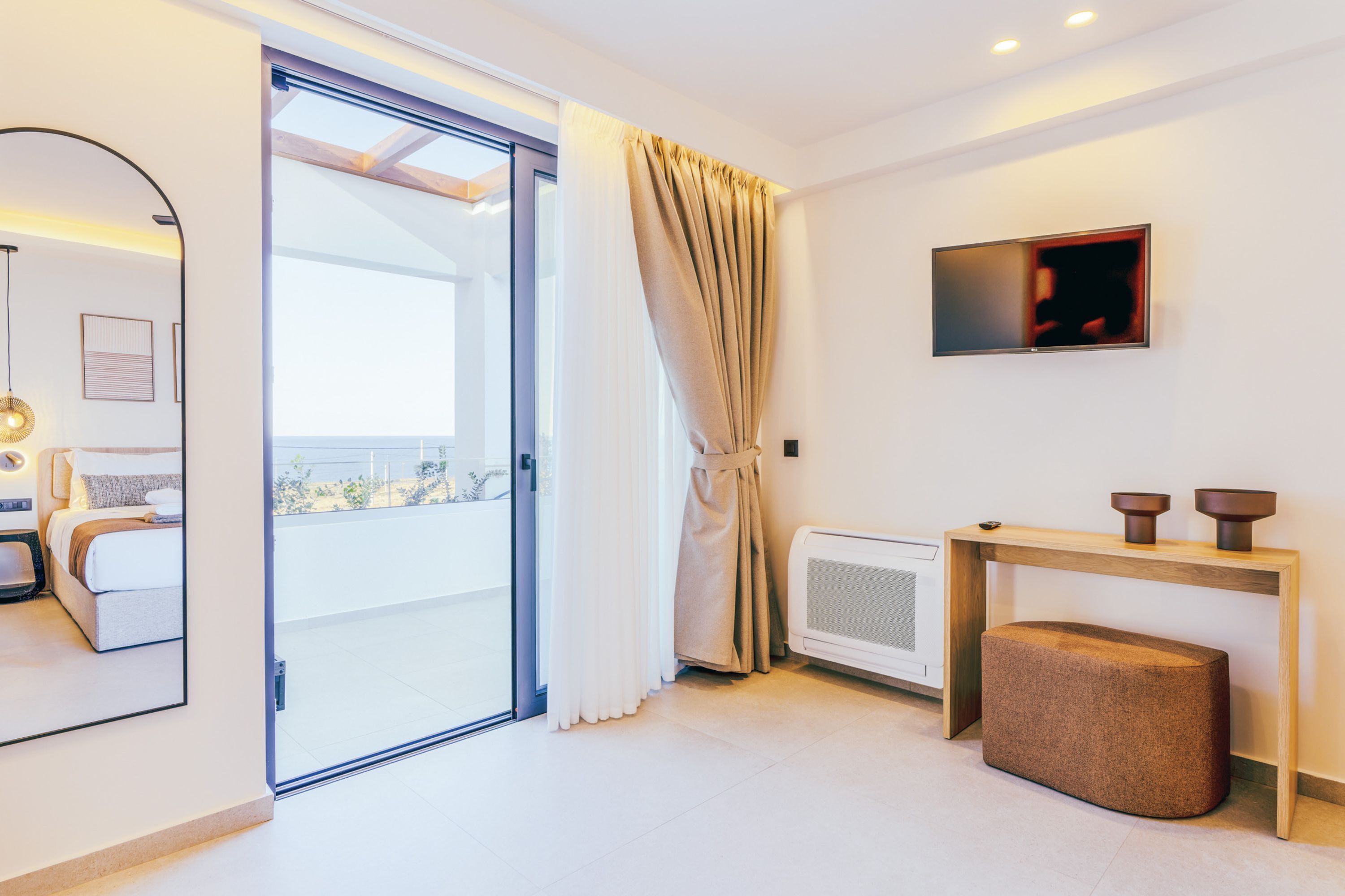 Een moderne hotelkamer met een schuifglasdeur naar een balkon, een bed, een nachtkastje met een lamp, en een klein zithoekje.