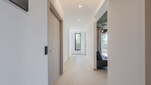 Un pasillo moderno con suelo de tonos claros, una puerta a la izquierda y una zona de estar con un banco y lámparas colgantes a la derecha.