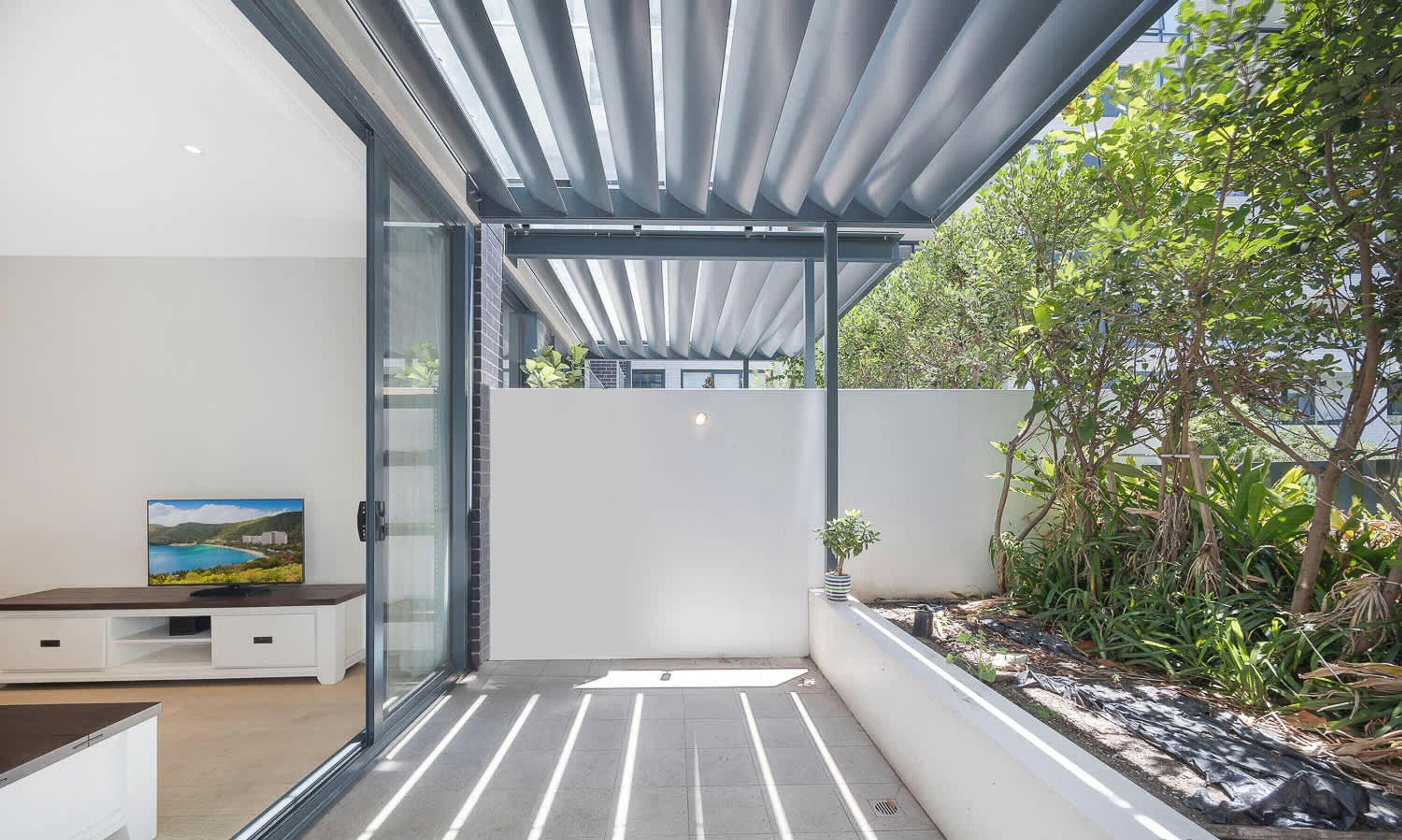A modern outdoor space with a glass door leading to a patio area, a white wall, and a view of greenery.