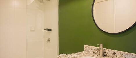 A bathroom with a glass shower enclosure, a sink with a granite countertop, and a round mirror.