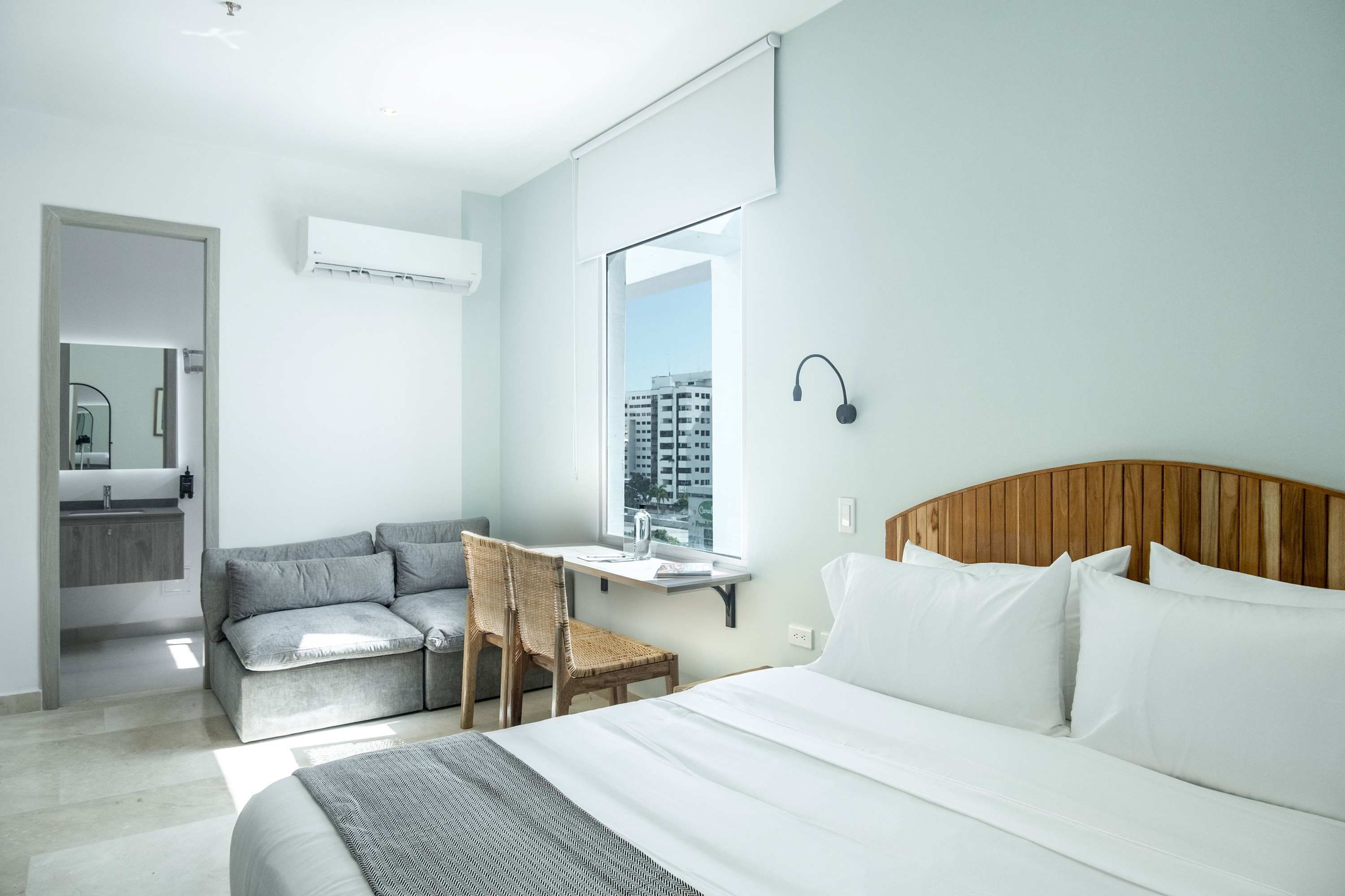 A modern hotel room with a bed, a sofa, a small table, and a window with a view of buildings.