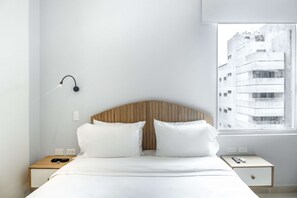 Ein schlicht eingerichtetes Hotelzimmer mit einem hölzernen Kopfteil, weißer Bettwäsche, einem Nachttisch mit Lampe und einem Fenster mit Blick auf die Stadt.
