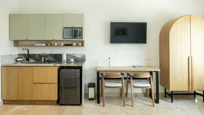 Eine kompakte Küche mit Mikrowelle, Spüle und kleinem Kühlschrank. Es gibt einen Holzkasten, einen Esstisch mit zwei Stühlen und ein an der Wand montiertes Fernsehgerät.