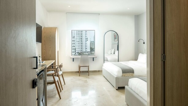 Ein modernes Hotelzimmer mit einem Bett, einem Schreibtisch mit Stuhl, einem Fenster mit Blick auf die Stadt und einem Badezimmer mit Spiegel und Waschbecken.