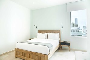 A modern bedroom with a wooden bed, white bedding, a nightstand, and a window with a city view. - w Trendy 2BR in the Heart of Bocagrande (Cartagena)