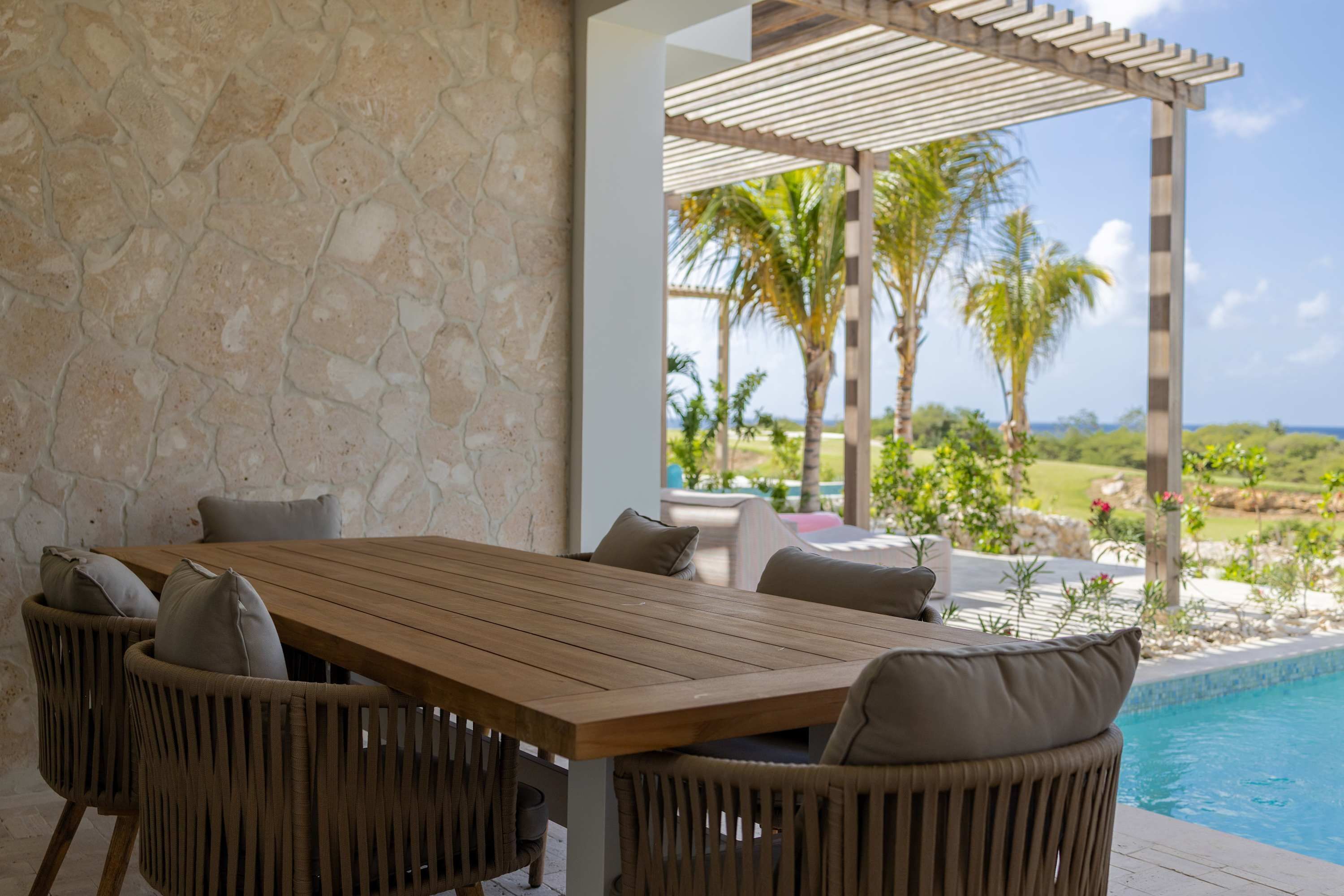 Mesa de madera con sillas de mimbre y cojines, frente a una pared de piedra con vistas a una piscina y paisaje tropical.
