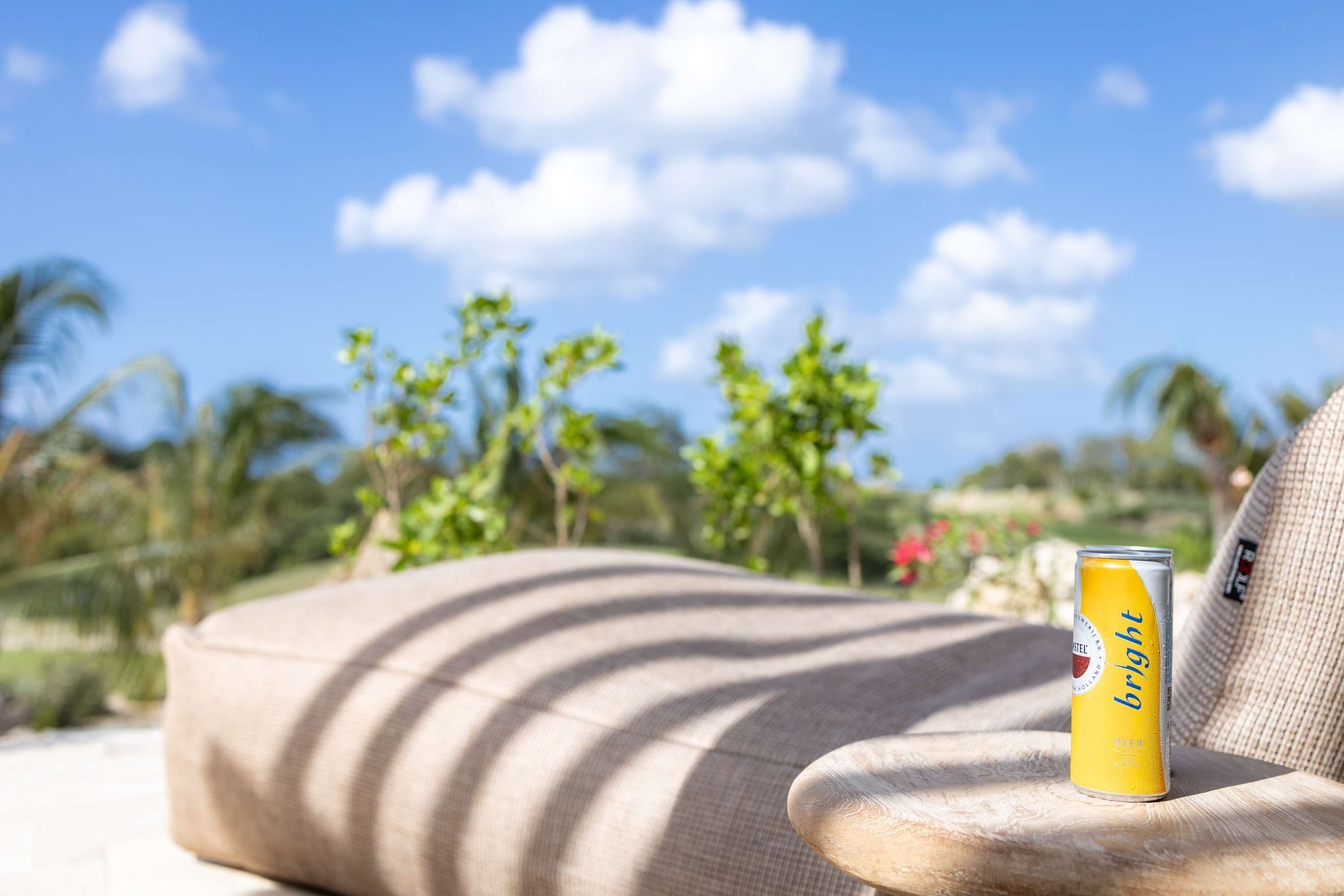Una lata de cerveza sobre un sofá color arena con vista a palmeras y un cielo despejado.