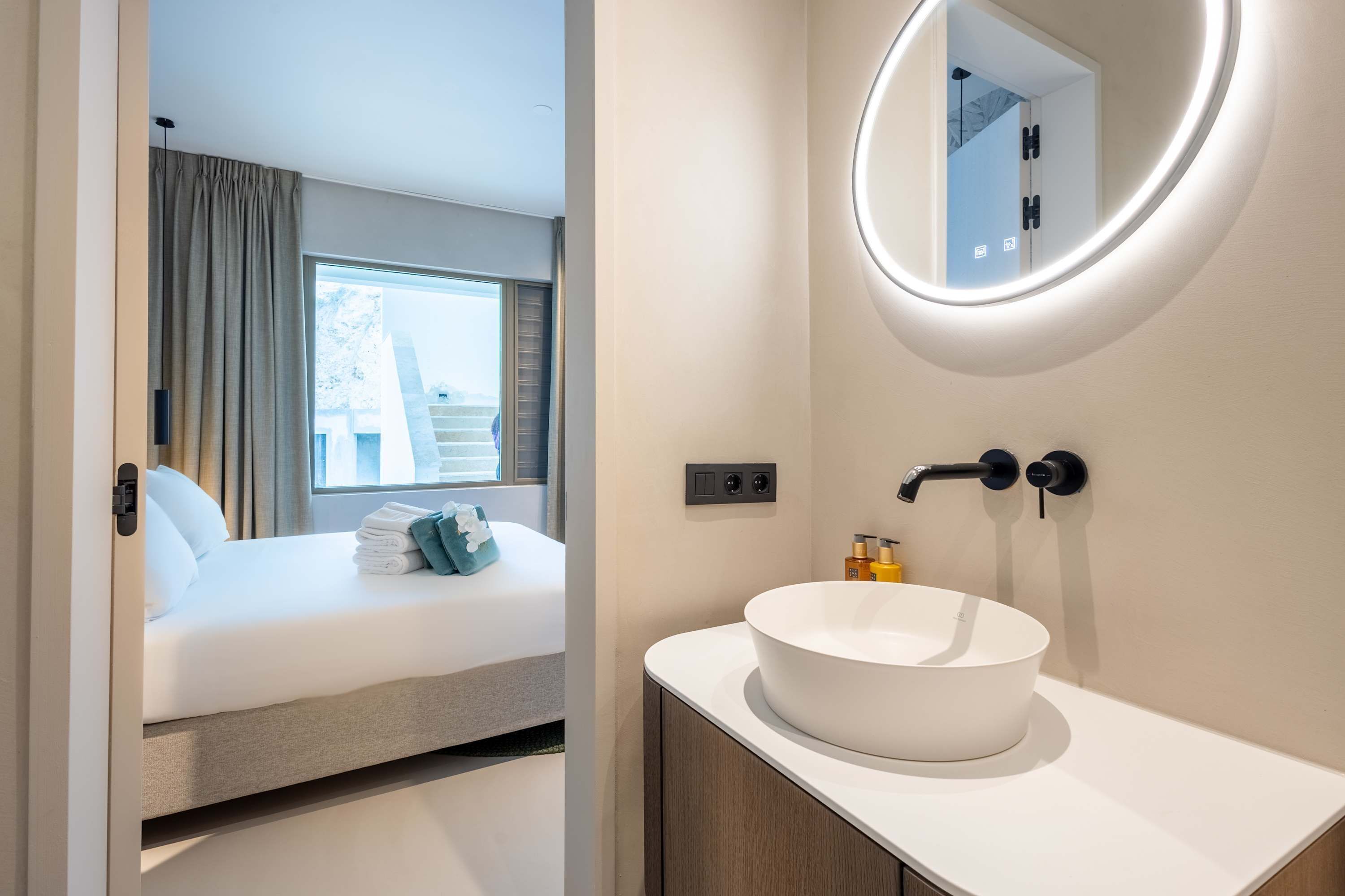 Una habitación de hotel moderna con cama, espejo redondo, lavamanos blanco y ventana con cortinas.