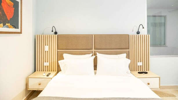Quarto de hotel moderno com uma cama grande, duas mesas de cabeceira com luminárias, uma janela e uma obra de arte emoldurada na parede.