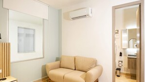 Quarto de hotel moderno com um sofá bege, um espelho grande, um aparelho de ar condicionado e vista para o banheiro por uma porta aberta.