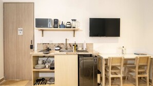 Cozinha compacta com micro-ondas, cafeteira, liquidificador e geladeira. Há uma TV na parede e um pequeno espaço de jantar com duas cadeiras.