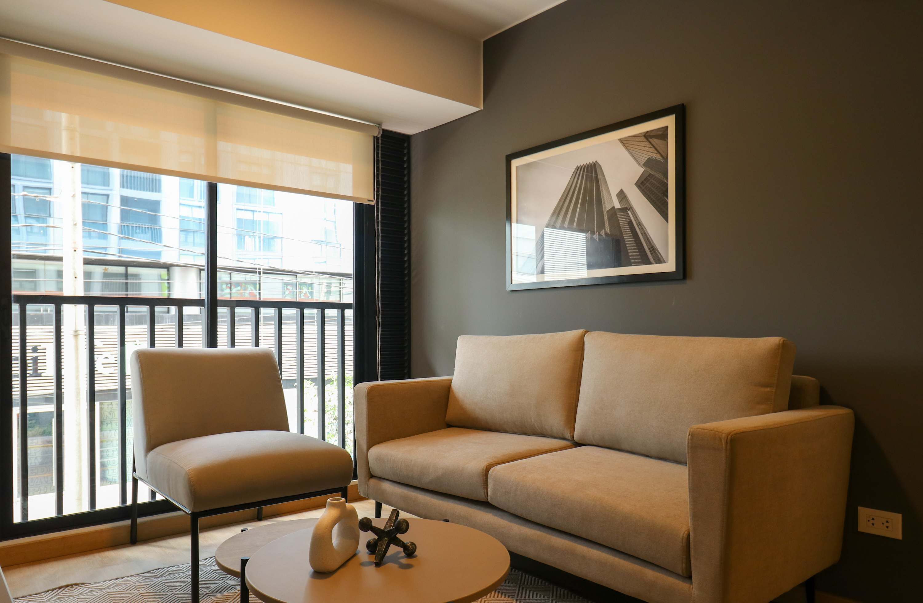 A modern living room with a beige sofa, a matching armchair, and a small coffee table. A framed picture hangs on the wall, and a window with blinds offers a view of buildings outside.