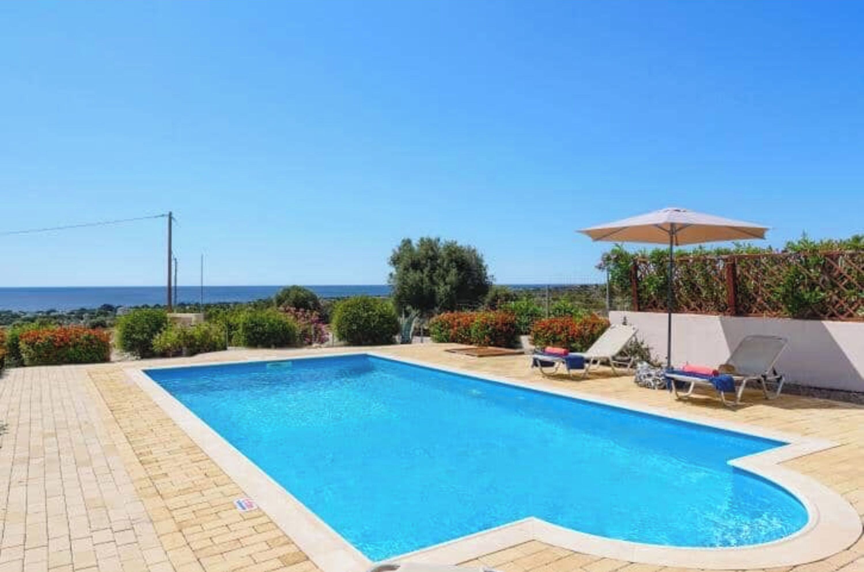 A rectangular swimming pool with a curved edge, surrounded by a tiled deck. There are lounge chairs and a sun umbrella near the pool.