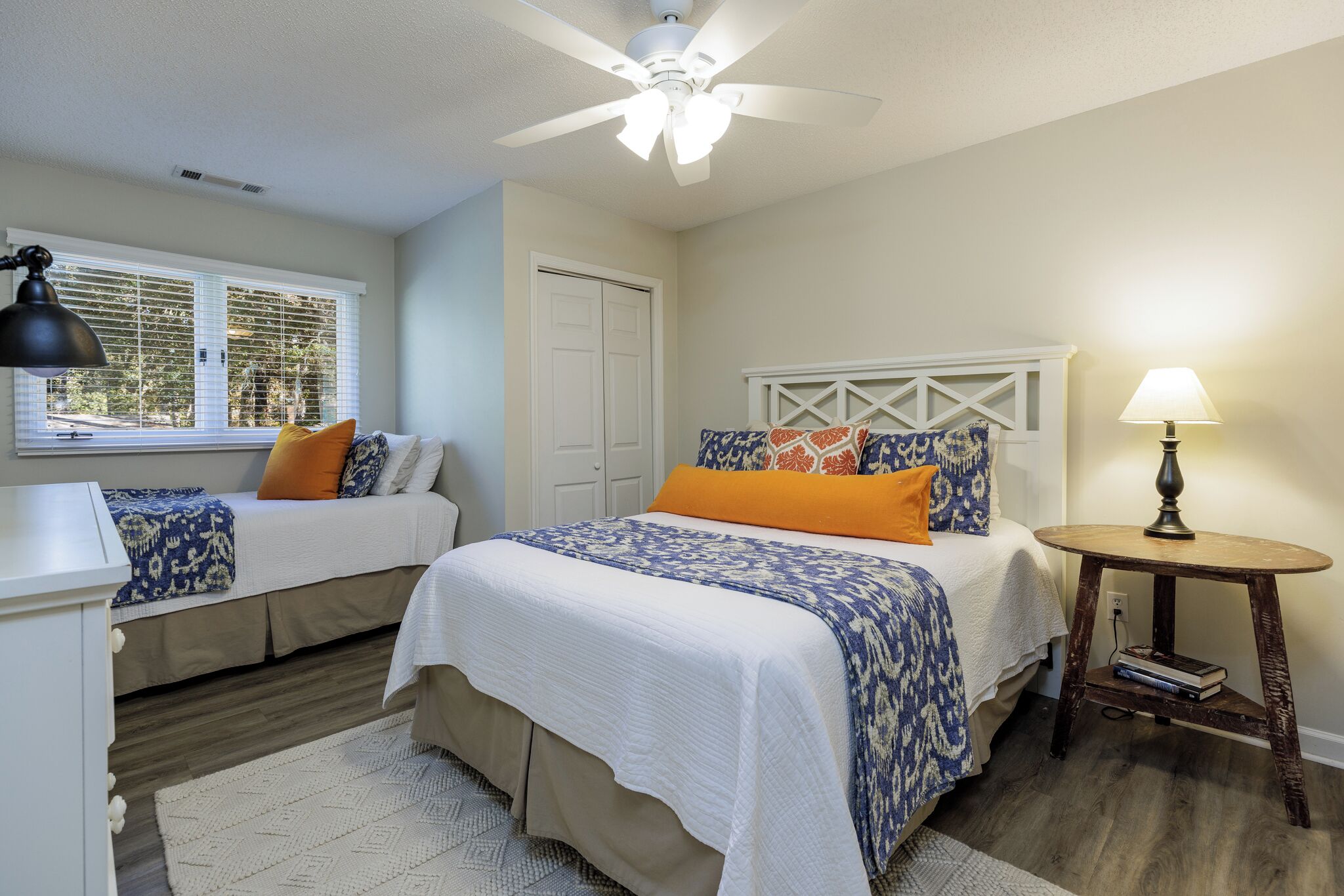 A bedroom with two beds, a ceiling fan, a bedside table with a lamp, and a window with blinds.