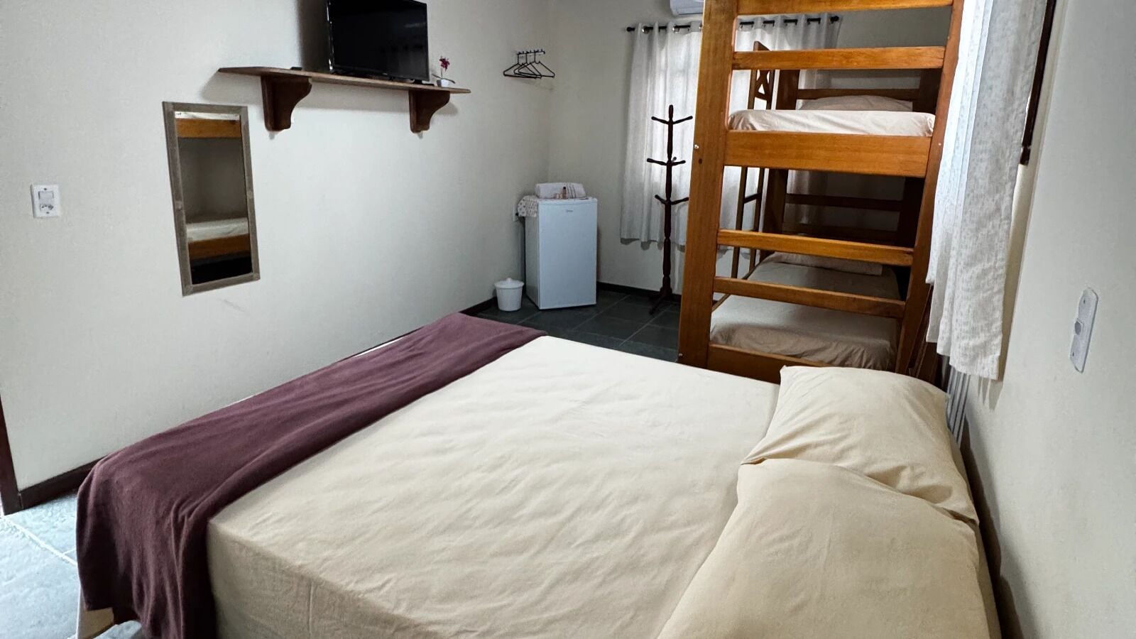 Una habitación pequeña y sencilla con una cama, una cama litera de madera, un espejo, un estante con un televisor y un electrodoméstico blanco pequeño.