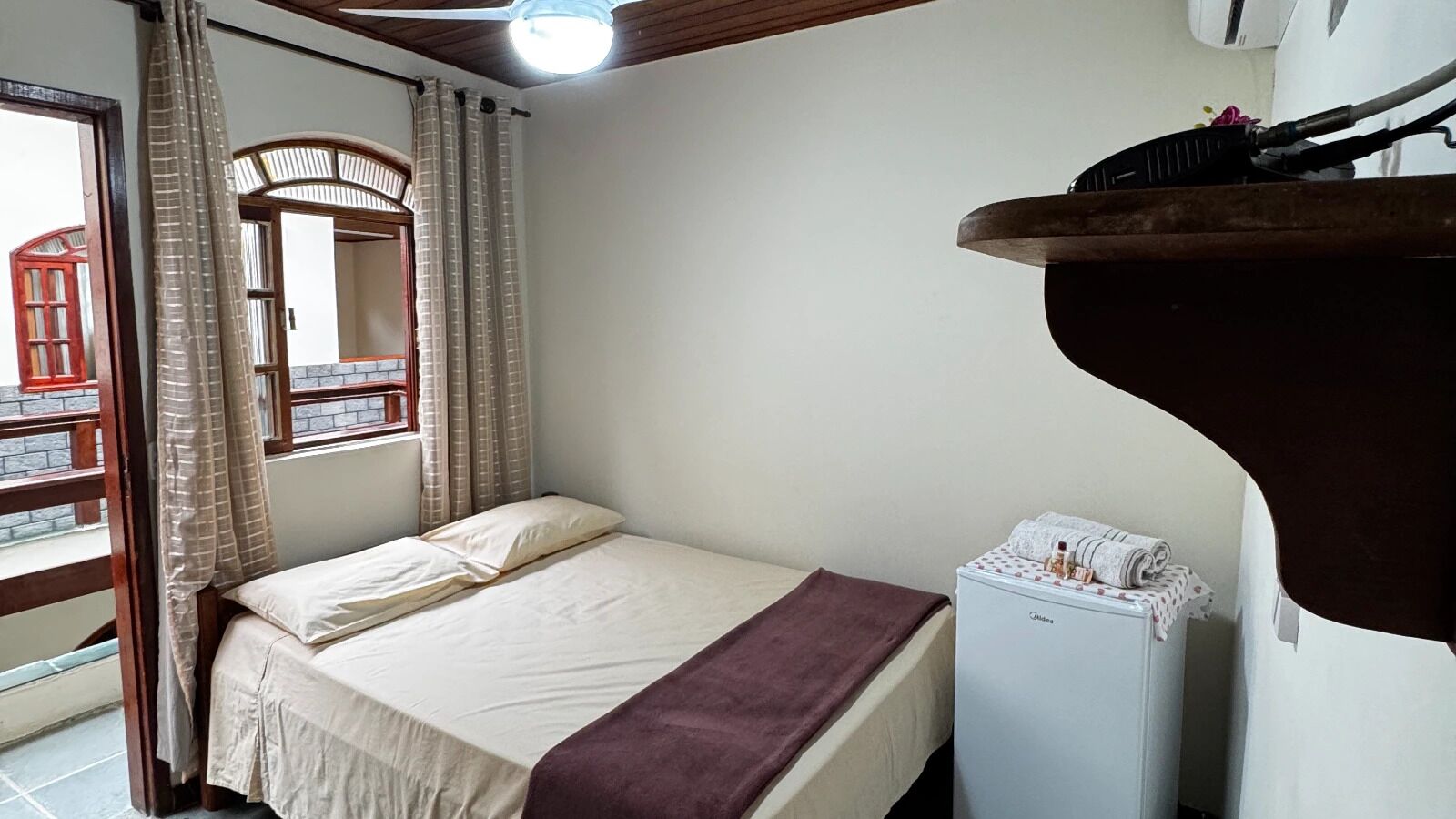 Un dormitori amb un llit, una finestra amb cortines, un ventilador de sostre i un aparell blanc petit.