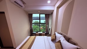 Quarto de hotel com uma cama grande, vista para área verde e um edifício pela janela, e uma pequena mesa com cadeira perto da janela.