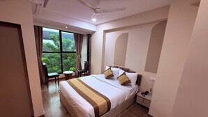 Quarto de hotel com uma cama grande, um ventilador de teto, vista para o verde pela janela e uma pequena mesa com cadeiras.