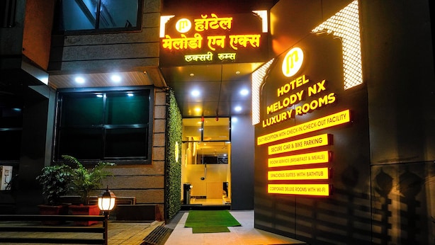 Entrada de hotel com placa indicando “Hotel Melody NX Luxury Rooms” e comodidades como recepção 24 horas, estacionamento seguro para carros e bicicletas, serviço de quarto, suítes espaçosas com banheira e quartos deluxe para empresas com Wi-Fi.