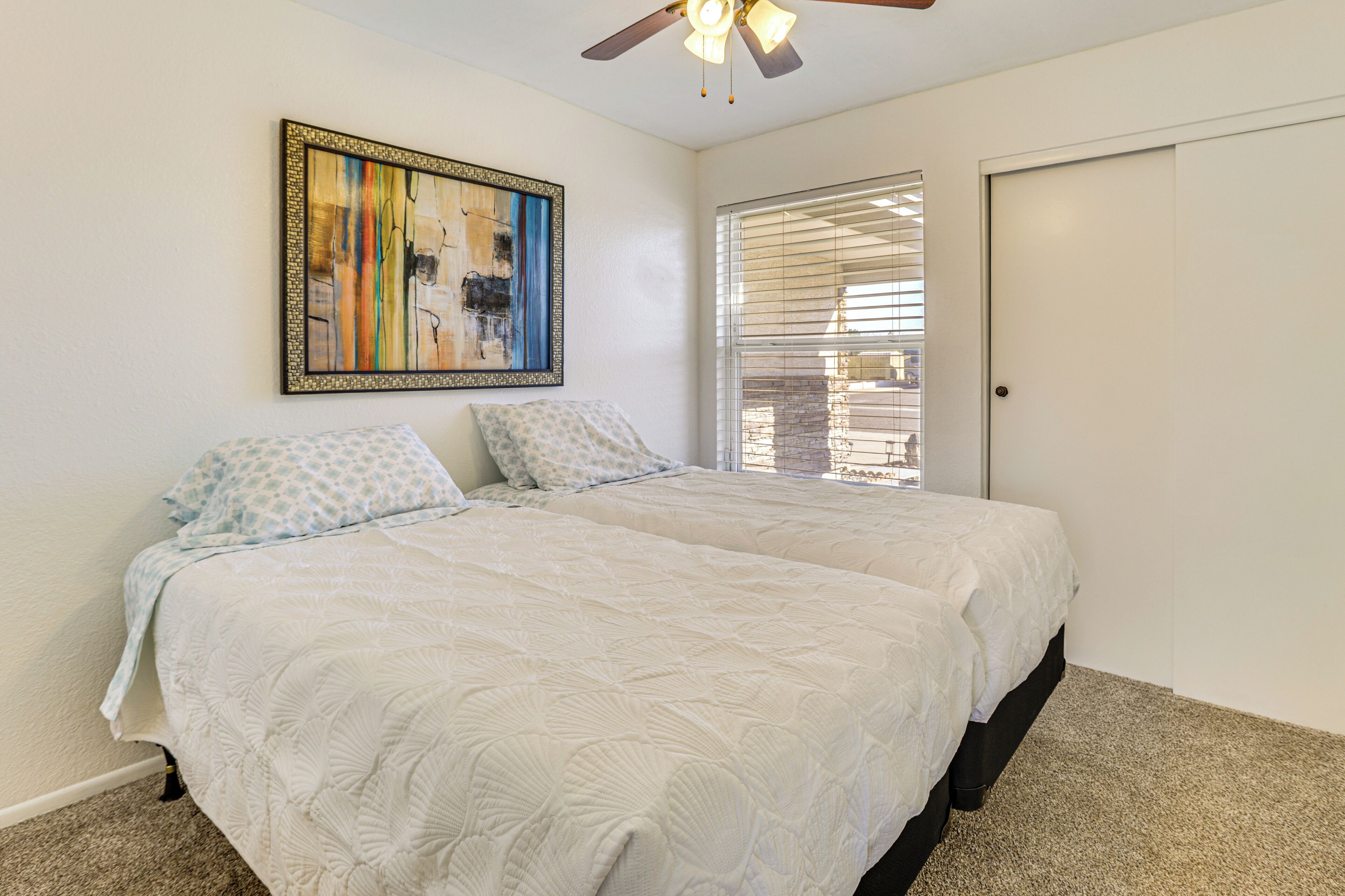 A bedroom with two beds, a ceiling fan, a window with blinds, and a framed abstract painting on the wall.