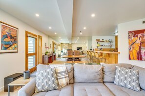 A modern living room with a grey sofa, patterned cushions, a small table, and a bench. A kitchen area with a dining table and chairs is visible in the background. - 180-degree Snow Park Views: Condo Near Deer Valley (Park City)