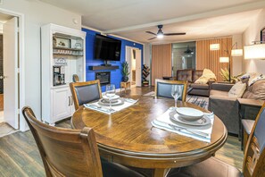 Ein Essbereich mit einem Holztisch, gedeckt für vier Personen, einem an der Wand befestigten Fernseher und einem Wohnzimmer mit Sofa und Deckenventilator.