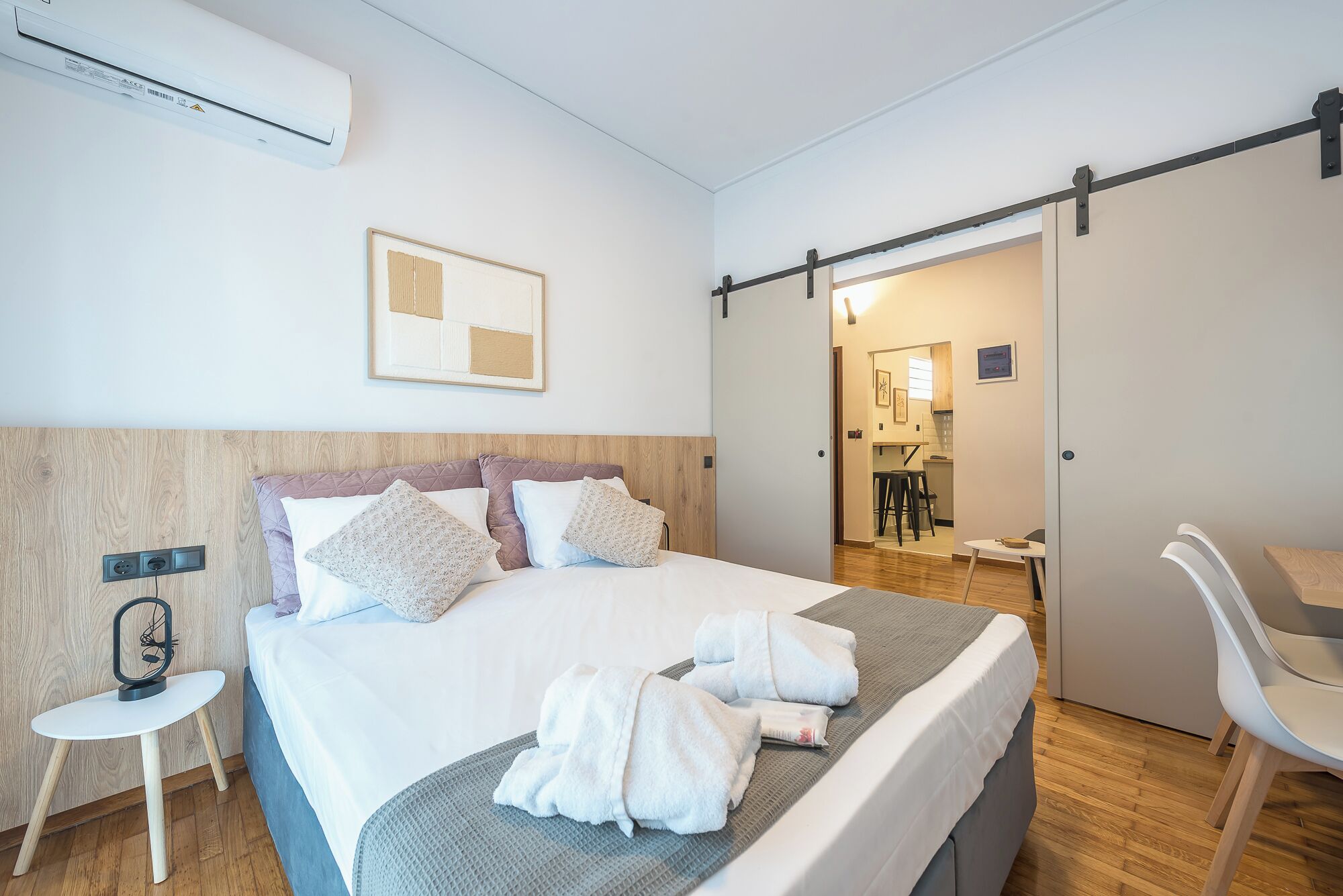 Hotellihuone, jossa on sänky, työpöytä, tuoli, yöpöytä, seinään asennettu ilmastointilaite ja ovi, joka johtaa kylpyhuoneeseen.
