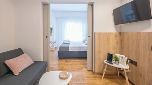 A modern hotel room with a sofa, a small table with a remote control, a TV, and a bed with a view to a balcony. - Prime City Stay by Cloudkeys (Athens)