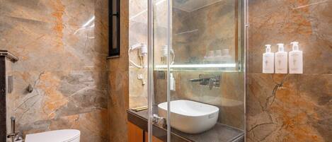 A bathroom with a marble wall, a glass-enclosed shower, a white sink, and toiletries on the wall.