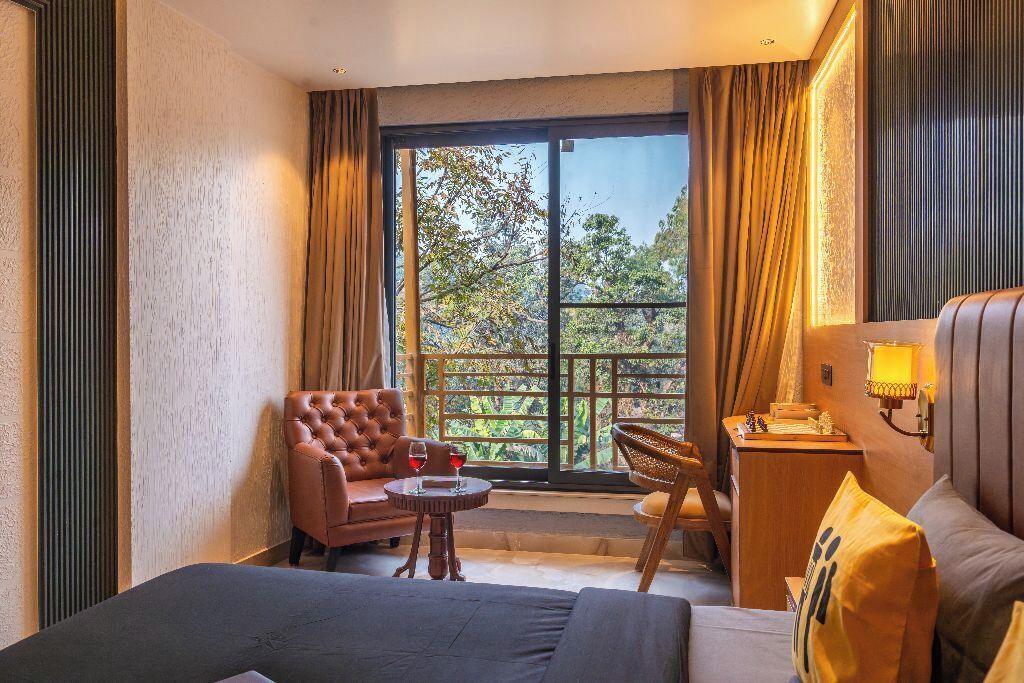 Une chambre d’hôtel avec un lit, un canapé, une chaise, une petite table avec un verre à vin, et une vue sur les arbres par une grande fenêtre.