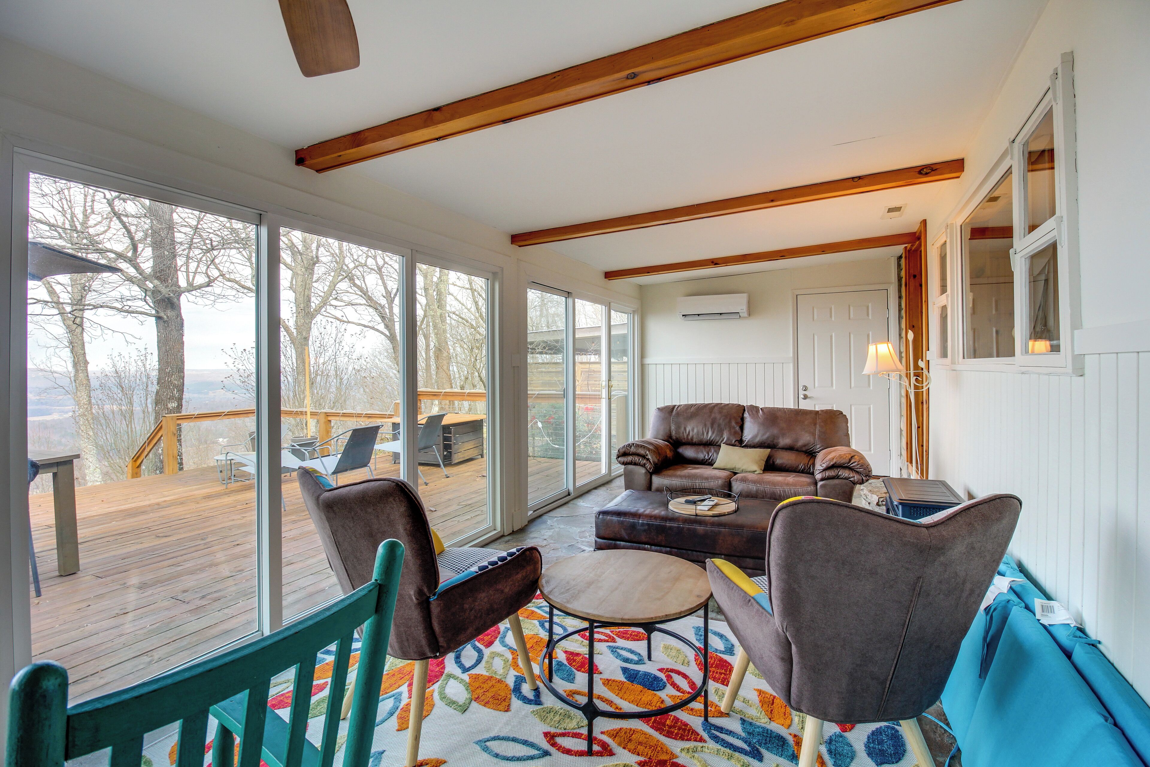 A spacious living area with a large window, a wooden ceiling, a brown leather sofa, and a colorful rug.