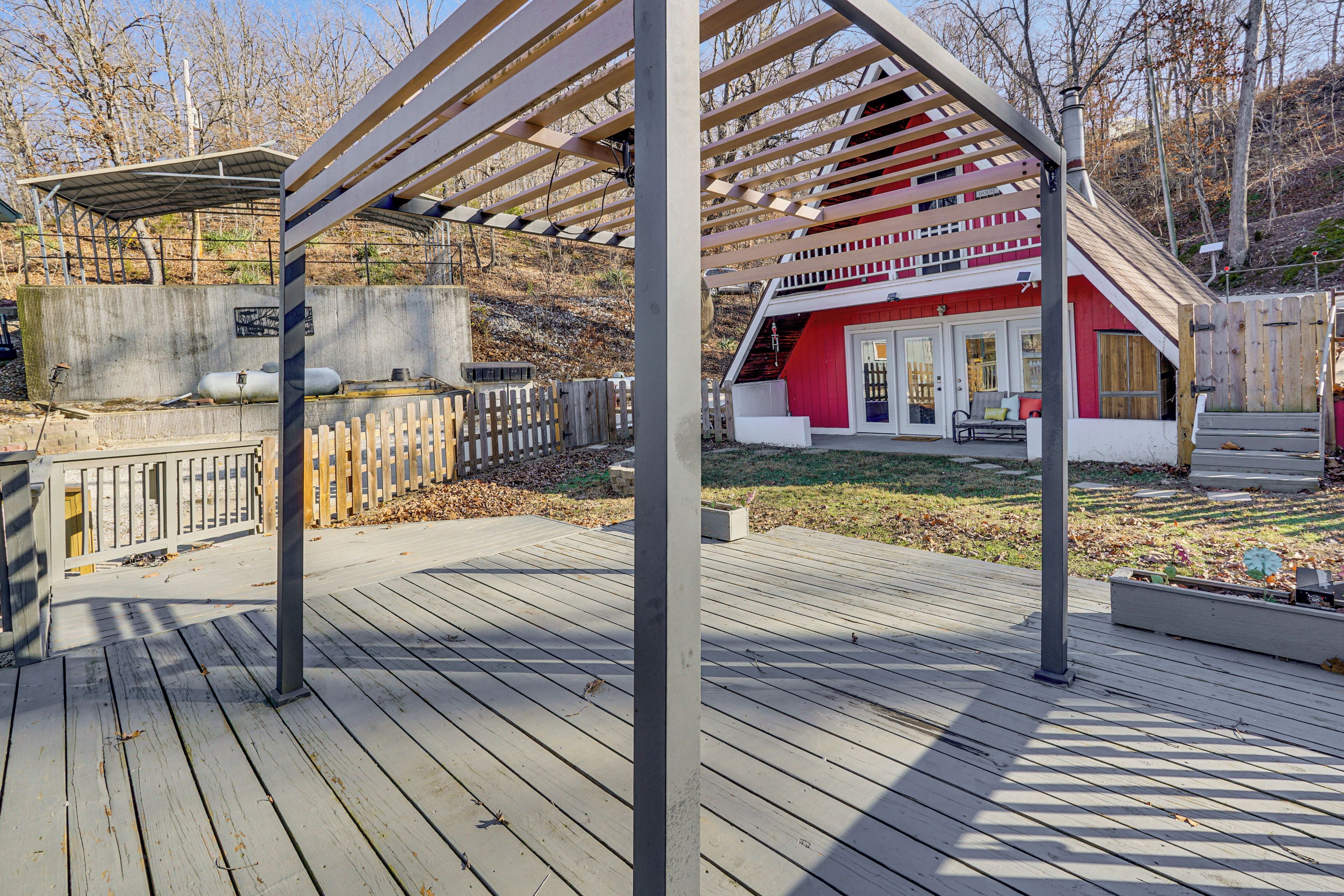 Una terraza de madera con una estructura de pérgola, un edificio rojo con puerta blanca y una pared de concreto con tanques.
