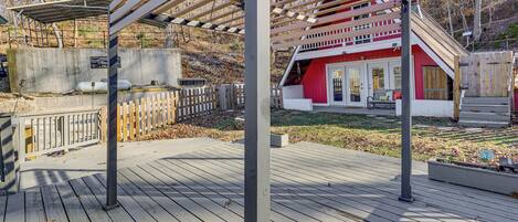 Una terraza de madera con una estructura de pérgola, un edificio rojo con puerta blanca y una pared de concreto con tanques.