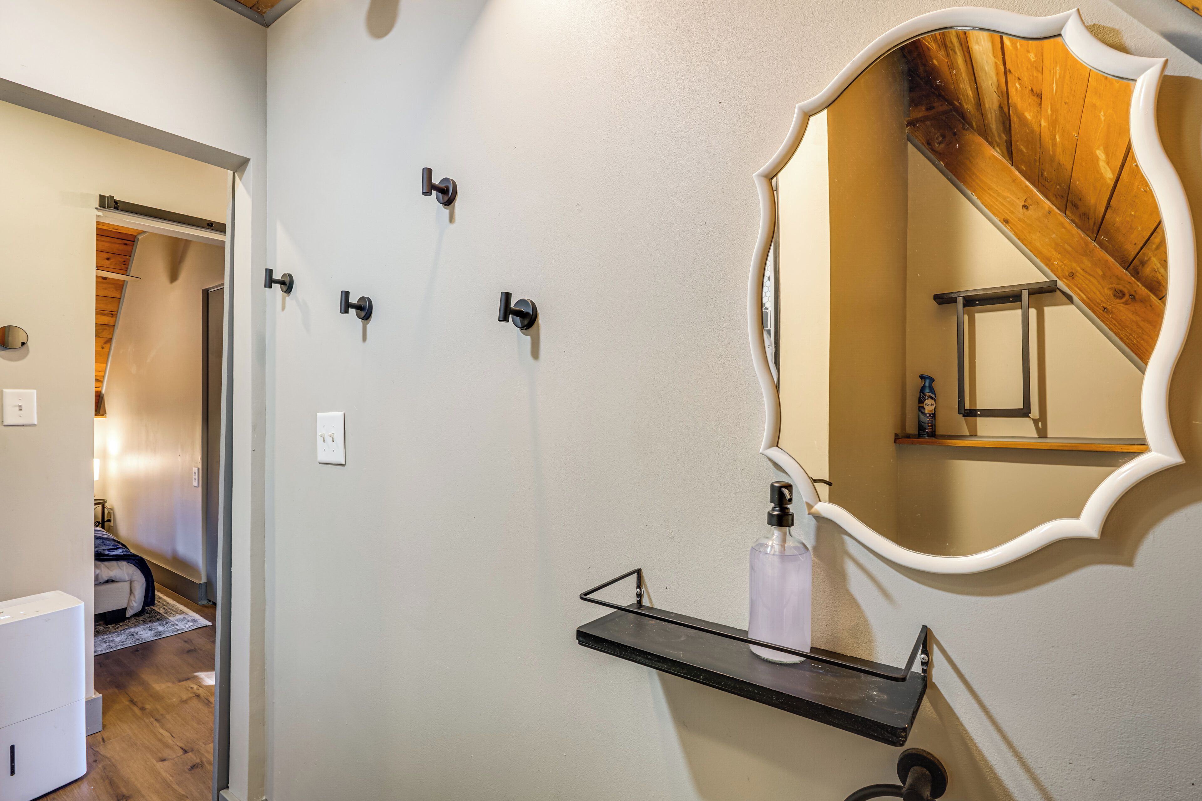 Ein Badezimmer mit Holzboden, einer weißen Wand und einem Spiegel mit dekorativem Rahmen. An der Wand sind drei schwarze Haken für Handtücher angebracht und es gibt ein schwarzes Regal mit einer Seifenspender.