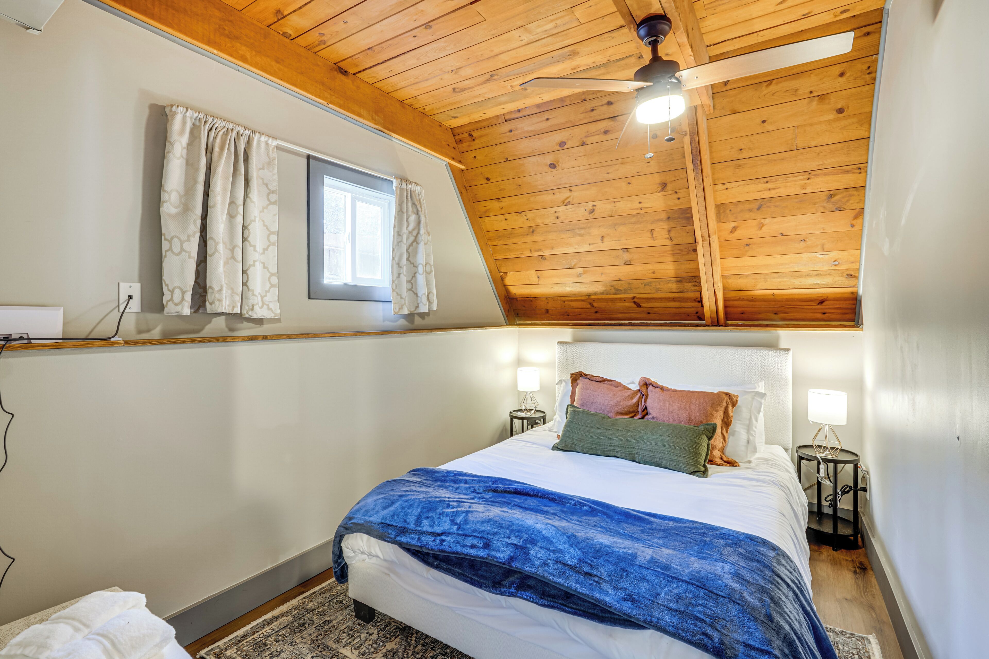 A bedroom with a sloped ceiling, a bed with a blue blanket, two bedside lamps, and a window with curtains.