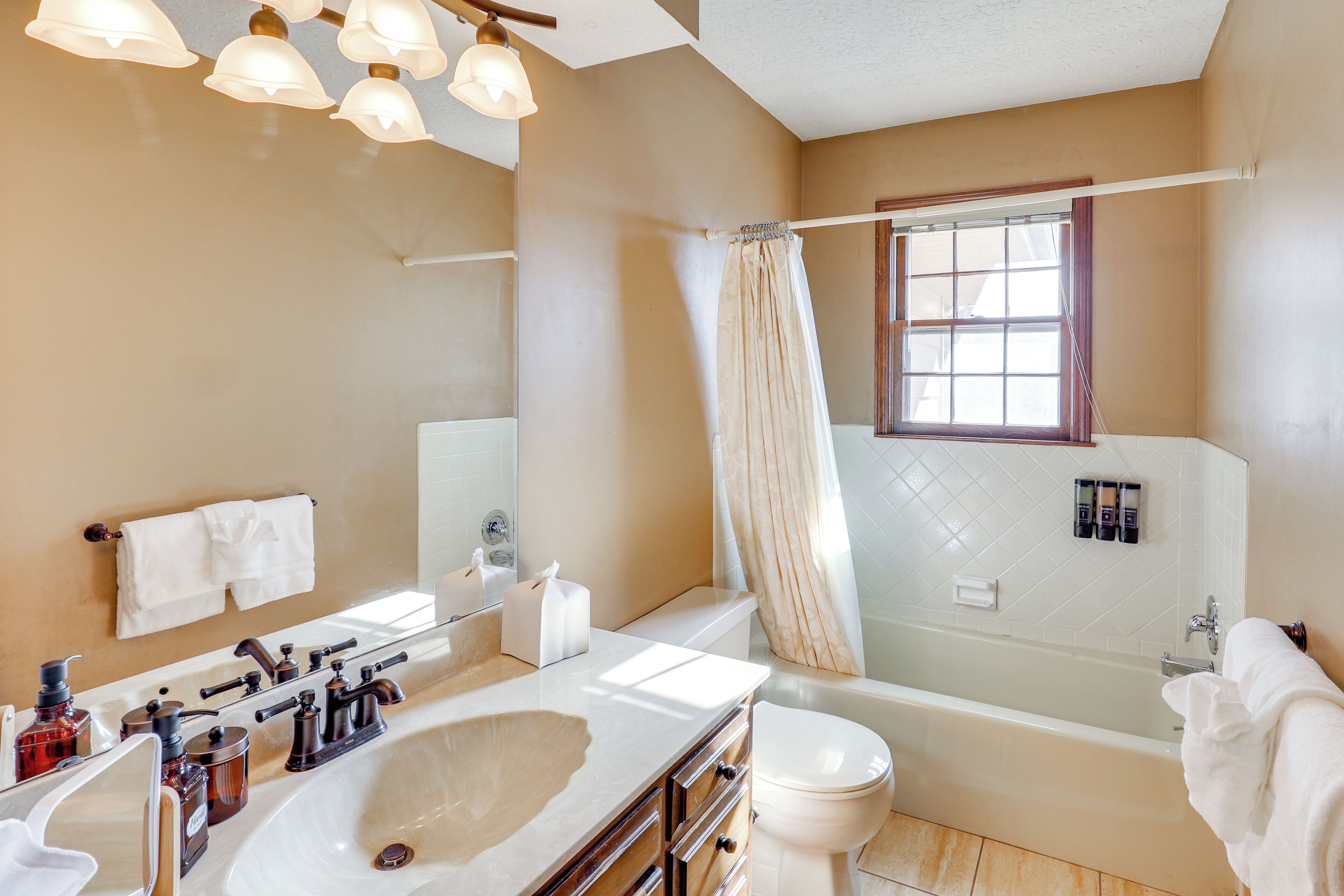 A bathroom with a large sink, mirror, and bathtub. There is a window with a curtain, and toiletries are visible on the counter.