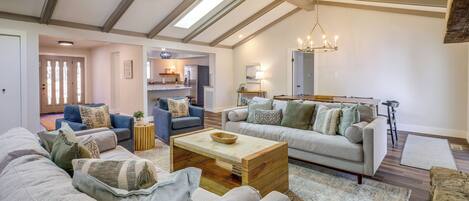 A spacious living room with a high ceiling, exposed beams, and a skylight. There are two sofas, a coffee table, and a dining area with a chandelier.