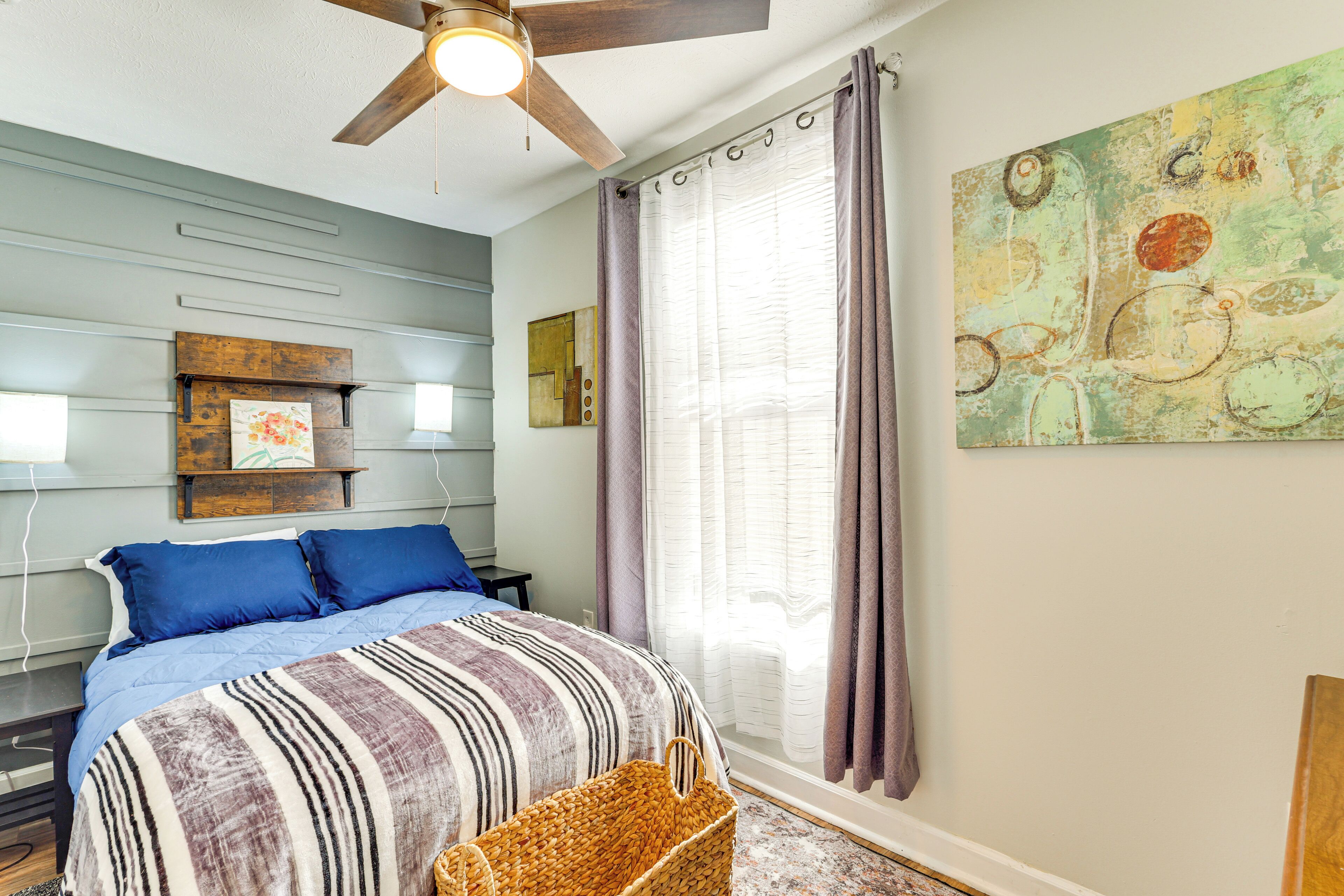 A bedroom with a bed, a ceiling fan, a window with curtains, and a wall-mounted artwork.