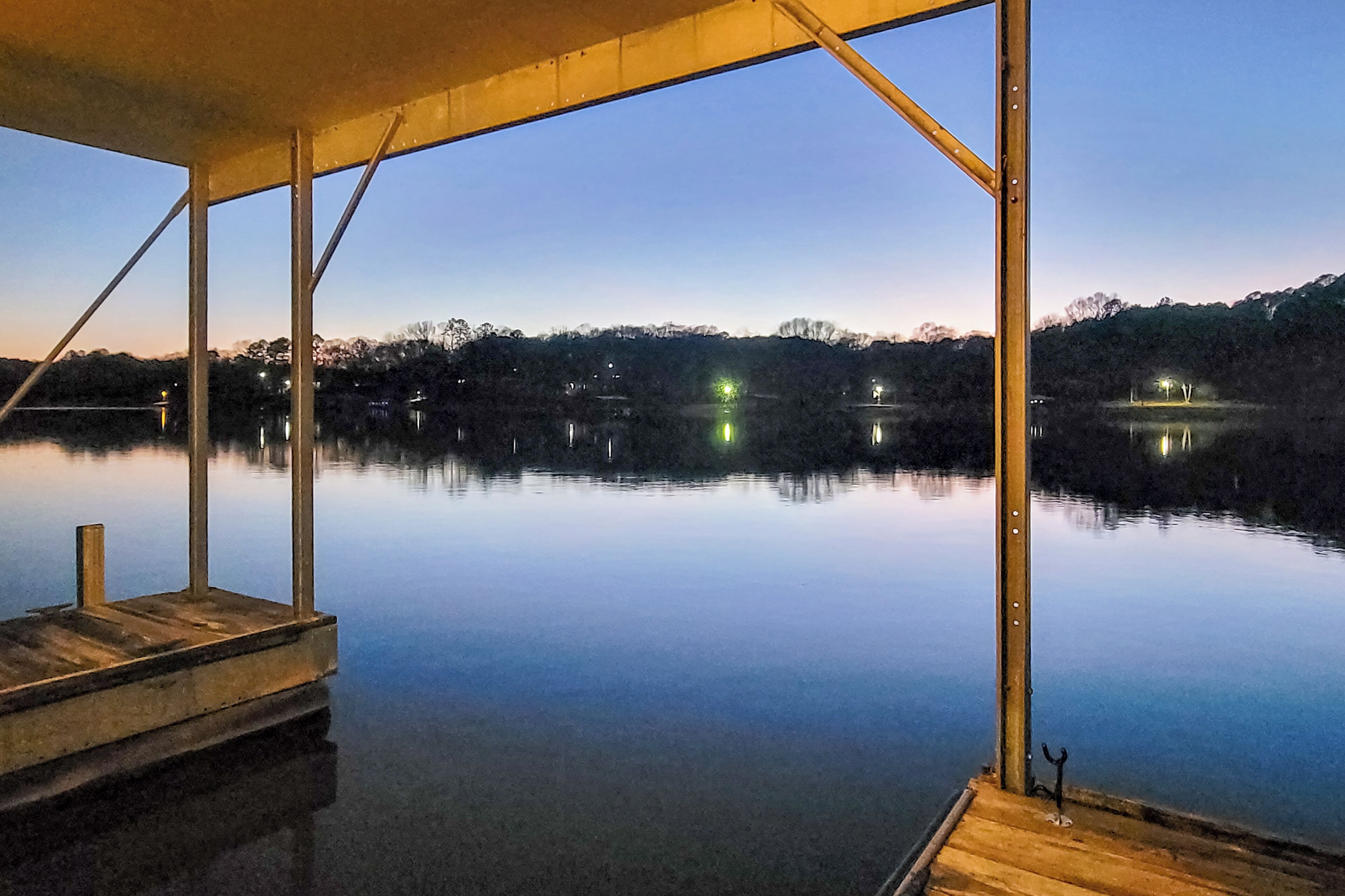A calm lake at dusk with a wooden dock and a metal structure overhead.