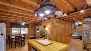 Interior de uma cabana de madeira com uma mesa de jantar, cadeiras, bancada de cozinha, ventilador de teto e uma parede com diversos avisos.
